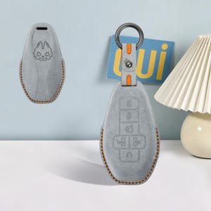 May include: A light gray key fob cover with orange stitching and a dark metal ring. The cover features a debossed graphic of a stylized wolf face on the front and molded button icons on the back. The material has a soft, suede-like texture.