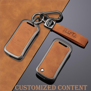 May include: Tan leather key fob covers with a gunmetal finish. One fob is attached to a key ring and a leather strap with the name "Elizabeth Taylor" printed on it. The bottom of the image has the text "CUSTOMIZED CONTENT".