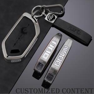 May include: A dark gray metal car key fob cover featuring a crescent moon and star design, paired with a matching black leather keychain strap embossed with the name Elizabeth Taylor and a circular charm showing a car silhouette icon.