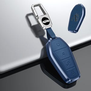 May include: A blue leather car key fob with a silver metal key ring. The fob has a textured surface and silver trim. The key ring has a car silhouette on a black background.
