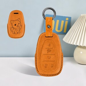 以下が含まれることがあります： ウィンクする子犬のイラストが型押しされたオレンジ色のレザー製カーキーフォブカバー。裏面にはボタンのアイコンが刻印されています。お揃いのレザーストラップとダークメタルの円形キーリングが付いています。
