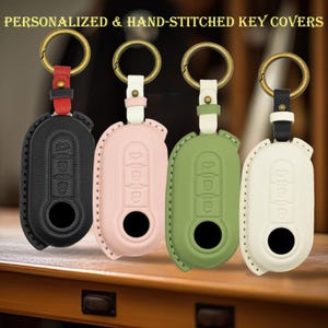 May include: Four personalized and hand-stitched key covers in black, light pink, sage green, and off-white. Each cover features a textured surface, embossed button icons, a circular cutout, and a brass-toned metal ring attached by a leather strap.