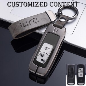 May include: A gunmetal grey key fob with a leather strap engraved with "Elizabeth Taylor." The fob has a black screen and white icons for keyless entry, a motorcycle, and a speaker. A silver key ring is attached.