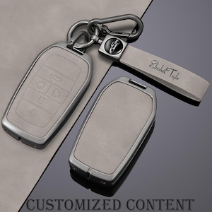 Puede incluir: Un juego de llaveros grises con marco de metal y correa a juego. La correa está grabada con el nombre "Elizabeth Taylor". Los llaveros tienen iconos de botones en relieve. Las palabras "CUSTOMIZED CONTENT" están en la parte inferior.