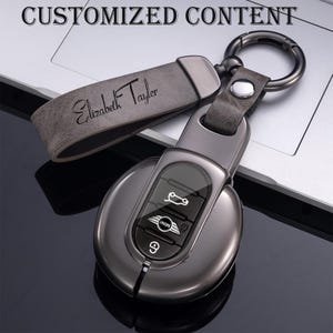 Può includere: Un portachiavi personalizzato con un cinturino in pelle grigia inciso con il nome "Elizabeth Taylor". Il portachiavi è attaccato a una chiave per auto in metallo grigio scuro. Il testo "CUSTOMIZED CONTENT" è in alto.