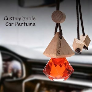 Könnte beinhalten: Ein hängender Auto-Lufterfrischer mit einem facettierten, rautenförmigen Glasflakon, der mit orangefarbener Flüssigkeit gefüllt ist. Er hat einen hellen Holzverschluss mit der Gravur Perfume und hängt an dunkelbraunen Kordeln mit Holzperlen.