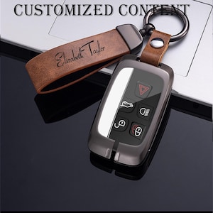 May include: A personalized car key fob with a brown leather strap and a metal key ring. The strap is engraved with the name "Elizabeth Taylor". The key fob is dark gray with white accents and black buttons.