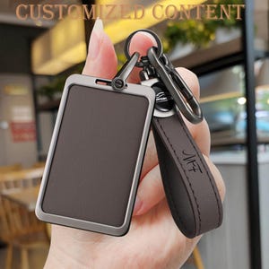 May include: A brown leather keychain with a rectangular card holder and a metal clasp. The keychain has a dark brown leather strap with the initials "NFT" embossed on it. The text "CUSTOMIZED CONTENT" is at the top.