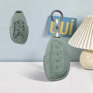 May include: A sage green suede car key fob cover featuring a detailed black line drawing of two wolves interacting. The cover includes a matching strap with a dark metal ring, white contrast stitching along the edges, and embossed button icons.