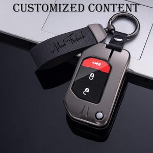 May include: A gunmetal gray car key fob with a black leather strap and a metal keyring. The key fob has red and black buttons. The leather strap is embossed with the name "Mark Frederick". The text "CUSTOMIZED CONTENT" is at the top of the image.