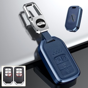 May include: A blue car key fob with a silver metal frame and a matching key cover. The fob has a leather texture and a silver car emblem. The key cover has the letter "A" and a leaf design.