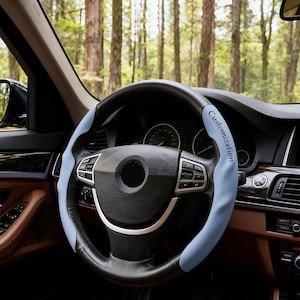 May include: A black steering wheel cover with light blue side panels and the word Customization printed in black on the right side. The cover is installed on a car steering wheel with silver accents and integrated control buttons.