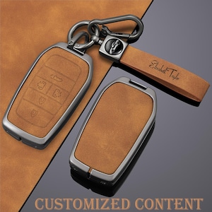 Puede incluir: Dos fundas de llavero de cuero marrón con marcos de metal plateado y una correa de cuero a juego. Una funda tiene símbolos de llave en relieve. La correa está grabada con "Elizabeth Taylor". Las palabras "CUSTOMIZED CONTENT" están en la parte inferior.