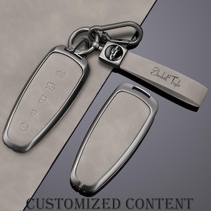 May include: A set of car key fobs and a keychain. The key fobs have a light gray suede-like finish with silver-tone metal trim. The keychain has a matching gray strap with the name "Elizabeth Taylor" and a car emblem.