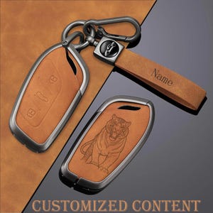 Puede incluir: Una funda de cuero color canela para llave de coche con marco de metal oscuro y una correa de llavero de cuero a juego con la palabra Name grabada. Una funda muestra una ilustración de tigre y la otra iconos de botones de llave de coche.