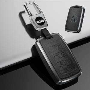 May include: Black leather car key fob with a silver metal frame and a silver key ring. The fob has several buttons and a car logo. A separate key fob is also visible with the letter "A".