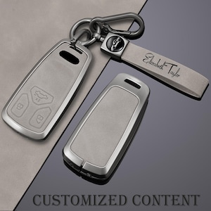 May include: A set of car key fobs with a gray leather-like finish and metallic silver trim. One fob is attached to a key ring and a gray leather strap with the name "Elizabeth Taylor" engraved. The image also includes the text "CUSTOMIZED CONTENT".