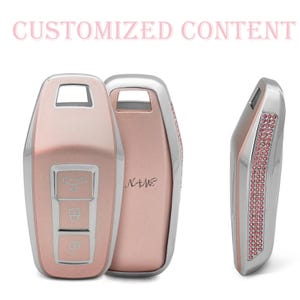 Pode incluir: Capa para chave de carro em ouro rosa com acabamento prateado e detalhes em strass rosa. A capa tem três botões e a palavra "NAME" está impressa na parte de trás. O texto "CUSTOMIZED CONTENT" está no topo.
