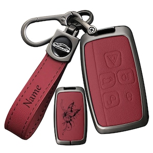 May include: A dark red leather car key fob cover set with gunmetal hardware. The set includes a strap with the word Name engraved, a small rectangular cover with a black butterfly graphic, and a larger cover with embossed vehicle control icons.