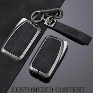May include: A set of car key fobs with a black leather and silver metal design. One fob has buttons, and the other is a solid rectangle. A black leather strap with the name "Elizabeth Taylor" is attached to a metal clasp.