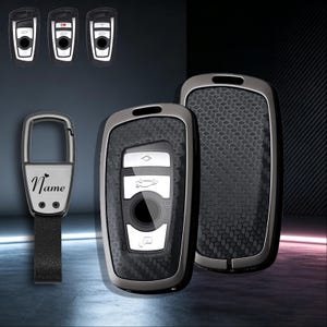 May include: Two car key fobs with a black carbon fiber pattern and metallic silver trim, alongside a metal keychain featuring a black leather strap and the word Name engraved on the front with a small heart symbol above the text.