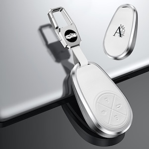 May include: A white leather car key fob with a silver metal frame and accents. The fob has four button icons and a matching key fob with the letter "A" and a floral design. A silver metal clip with a car icon is attached.