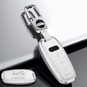 May include: A white and silver car key fob with a matching keychain. The fob has three buttons and a leather-like texture. The keychain features a car emblem and a silver clasp. A separate silver tag is engraved with a name and date.