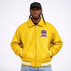 Puede incluir: Una chaqueta de cuero amarillo brillante con cremallera, cuello y pu&ntilde;os y dobladillo a rayas blancas. La chaqueta presenta un parche en el pecho con el texto "NIREX" y un dise&ntilde;o patri&oacute;tico. La persona lleva una gorra de b&eacute;isbol negra.