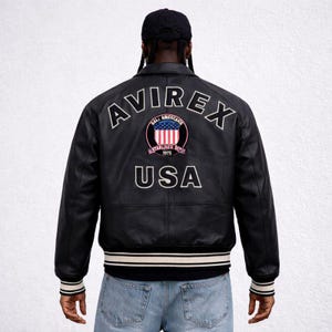 Puede incluir: Chaqueta de cuero negra con pu&ntilde;os y dobladillo a rayas blancas y negras. La parte trasera presenta la palabra "AVIREX" sobre un emblema circular con un dise&ntilde;o rojo, blanco y azul y la palabra "USA" debajo.