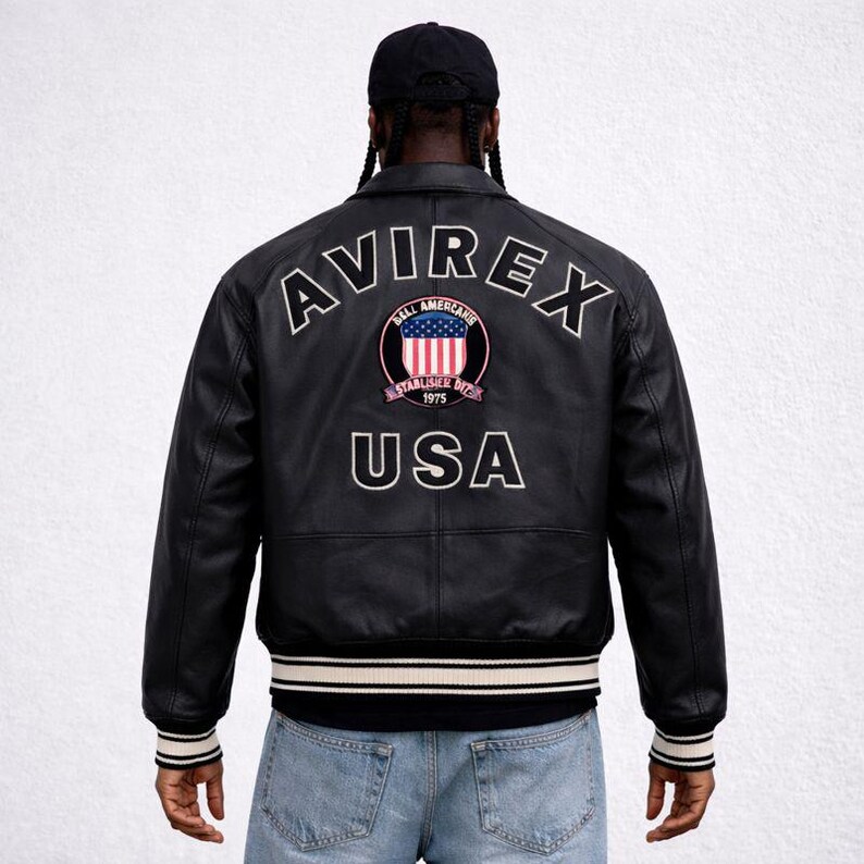 Puede incluir: Chaqueta de cuero negra con pu&ntilde;os y dobladillo a rayas blancas y negras. La parte trasera presenta la palabra "AVIREX" sobre un emblema circular con un dise&ntilde;o rojo, blanco y azul y la palabra "USA" debajo.