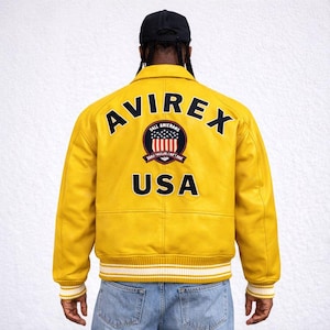 Puede incluir: Chaqueta de cuero amarilla con la palabra "AVIREX" en la espalda. Debajo, un emblema circular con las palabras "BALL AMIERNAS" y "USA". La chaqueta tiene pu&ntilde;os a rayas blancas y amarillas y un cuello negro.