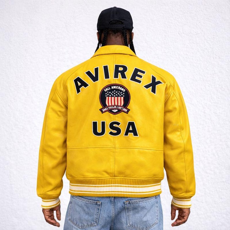Puede incluir: Chaqueta de cuero amarilla con la palabra "AVIREX" en la espalda. Debajo, un emblema circular con las palabras "BALL AMIERNAS" y "USA". La chaqueta tiene pu&ntilde;os a rayas blancas y amarillas y un cuello negro.