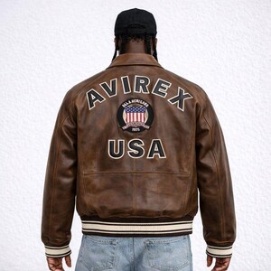 Puede incluir: Chaqueta bomber de cuero marr&oacute;n con la palabra "AVIREX" en la espalda. Un parche con la bandera americana y la palabra "USA" est&aacute; debajo del nombre de la marca. La chaqueta tiene cuello y pu&ntilde;os acanalados con rayas blancas.