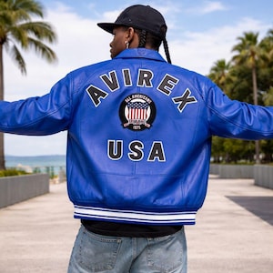 Puede incluir: Chaqueta de cuero azul brillante con la palabra "AVIREX" arqueada sobre los hombros. Debajo, un parche circular con las palabras "ALL AMERICANS" y "USA". La chaqueta tiene un ribete a rayas blancas y azules.