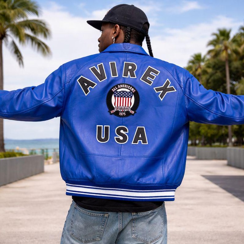 Puede incluir: Chaqueta de cuero azul brillante con la palabra "AVIREX" arqueada sobre los hombros. Debajo, un parche circular con las palabras "ALL AMERICANS" y "USA". La chaqueta tiene un ribete a rayas blancas y azules.