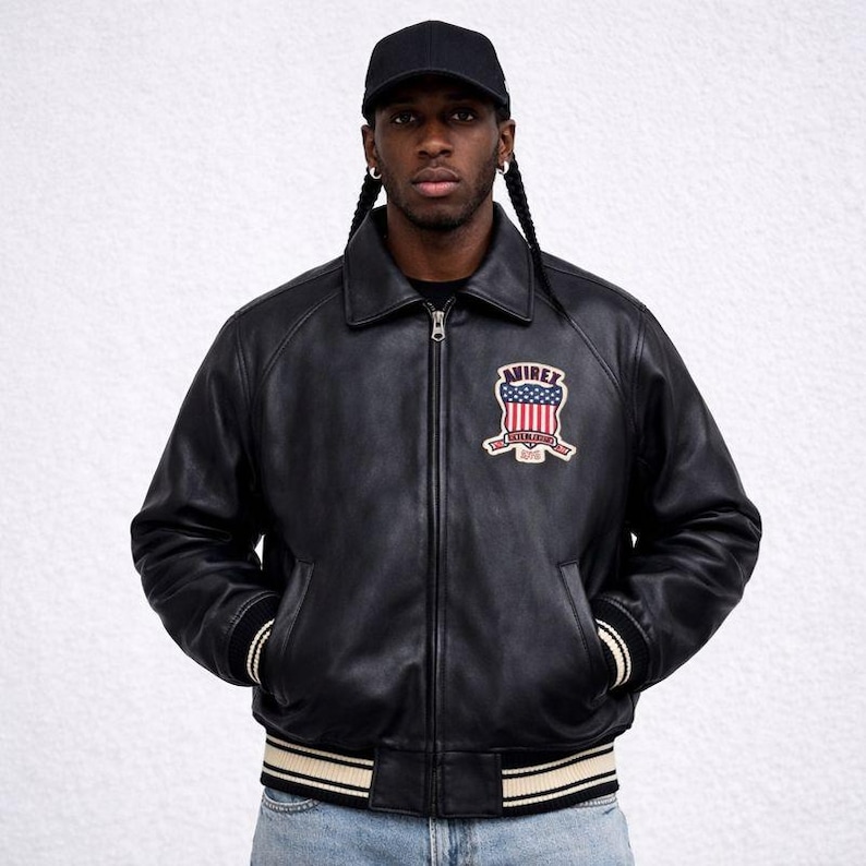 Puede incluir: Chaqueta de cuero negra con cuello, cierre de cremallera y parche en el pecho. El parche presenta un dise&ntilde;o rojo, blanco y azul con el texto "AVIREX ESTABLISHED 1975". La chaqueta tiene pu&ntilde;os y cintura a rayas.