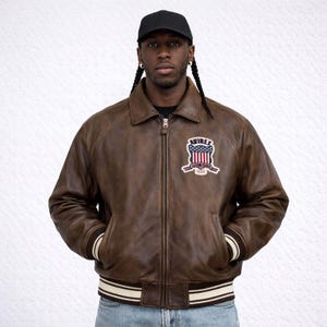 Puede incluir: Chaqueta bomber de cuero marr&oacute;n con cuello, cierre de cremallera y pu&ntilde;os y dobladillo a rayas. La chaqueta presenta un parche en el pecho con el texto "AVIREX" y un dise&ntilde;o de bandera americana. La persona en la imagen lleva una gorra de b&eacute;isbol negra.
