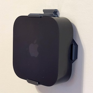 May include: A black Apple TV device mounted on a wall with a custom-fit, dark gray bracket. The Apple logo is visible on the front of the device. The bracket securely holds the device against the wall.