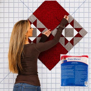 May include: A white design wall with a blue grid pattern, featuring a partially assembled quilt block made of dark red and grey patterned fabric squares. A plastic product package labelled Fons & Porter Design Wall is positioned in the lower right corner.