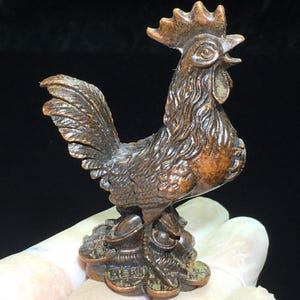 May include: A small, dark bronze-toned metal figurine of a rooster standing atop a cluster of traditional Chinese ingots and circular coins. The rooster features detailed plumage, a prominent comb, and an open beak, with a textured, weathered finish.