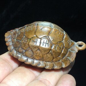 Puede incluir: Un colgante con forma de caparazón de tortuga marrón y texturizado con un acabado liso y pulido. El caparazón presenta una serie de marcas grabadas, geométricas y similares a caracteres en su superficie. Tiene una pequeña anilla circular.