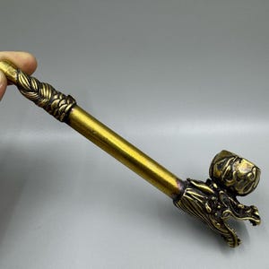May include: A metallic gold-toned smoking pipe featuring an elongated cylindrical stem with a braided texture at the mouthpiece end. The bowl end is shaped like a detailed dragon head with an open mouth, showcasing intricate scales and relief carvings.