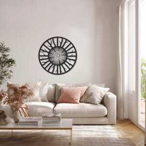 May include: A black, circular metal wall art piece resembling a cross-section of a tree trunk. The design features concentric circles and radiating branches. The artwork is mounted on a white wall above a cream-colored sofa with decorative pillows.