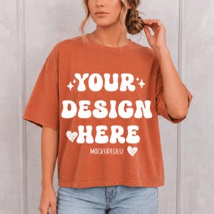 May include: A rust-colored short-sleeve t-shirt featuring the white text YOUR DESIGN HERE in a bold, wavy font. The design includes two small white heart icons and the word MOCKUPLULU printed in a smaller, simple white font below the main text.