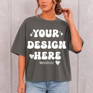 May include: A charcoal gray short-sleeve t-shirt featuring the white text "YOUR DESIGN HERE" in a bold, rounded font. The design includes two small star icons and two heart icons, with the text "MOCKUPLULU" printed in a smaller font below.