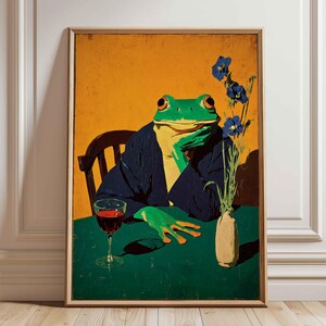 May include: A framed wall art print featuring a green frog with orange eyes wearing a dark blue jacket, sitting at a table with a glass of red wine and a small vase containing blue flowers against a solid mustard yellow background.