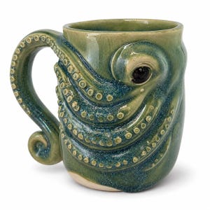 Peut inclure: Une tasse en céramique avec un vernis brillant vert et bleu, présentant un motif de pieuvre sculpté. L'anse est en forme de tentacule, et le corps de la tasse affiche des tentacules en relief texturés avec des ventouses et un œil sombre.