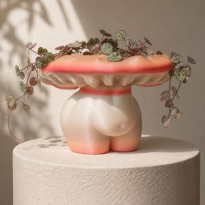 May include: A unique, mushroom-shaped planter with a white body and a pink and orange top. The planter has a decorative pink stripe around the middle and is filled with trailing green and purple plants. The planter is sitting on a round, light-colored pedestal.