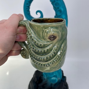 May include: A green ceramic mug with an octopus design. The mug features a tentacle-shaped handle and a textured surface with suction cup details. A teal octopus tentacle is behind the mug, and the mug is on a black base.
