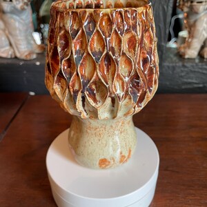 Può includere: Un vaso in ceramica con un motivo a nido d'ape strutturato nei toni del marrone e del beige. Il vaso ha una parte superiore ampia e una base arrotondata, appoggiato su un piedistallo bianco. Il design generale è organico e artistico.